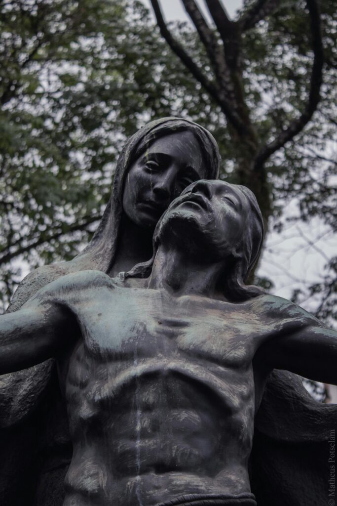 Perdas e Lutos - Estátua de bronze retratando figura masculina caída nos braços de figura feminina, em expressão de dor e acolhimento, com árvores ao fundo.