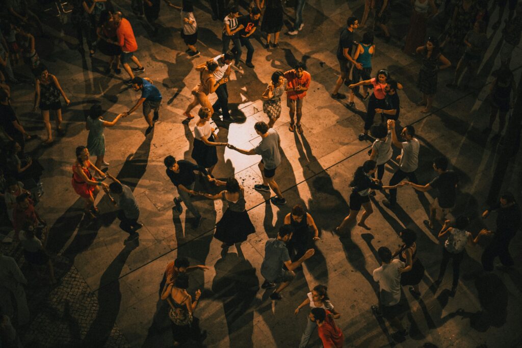 Grupo de pessoas dançando em círculo em uma praça à noite, com iluminação dramática projetando longas sombras no chão.