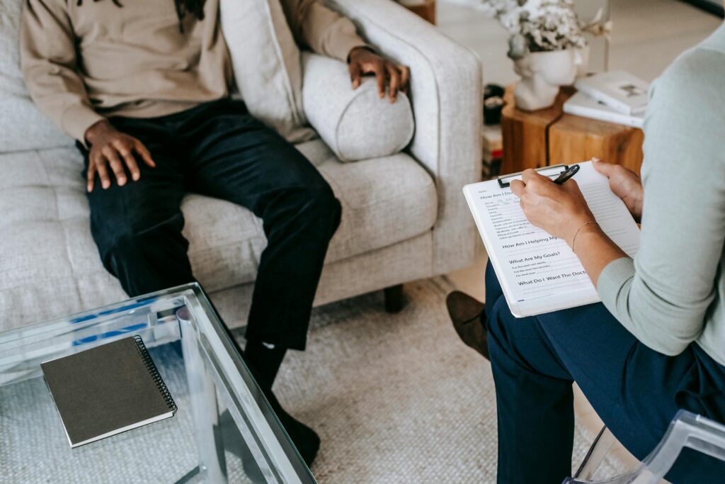 Pessoa em sessão de terapia sentada em sofá claro, enquanto terapeuta faz anotações em prancheta, em ambiente moderno e acolhedor.