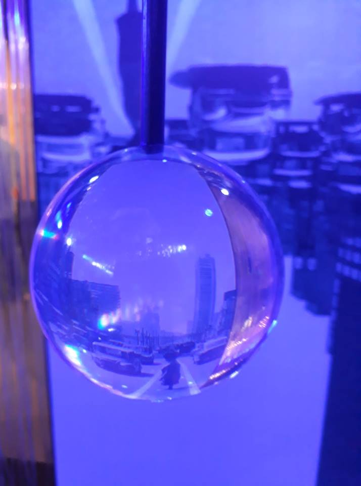 Imagem de uma esfera translúcida pendurada refletindo uma avenida urbana com prédios e veículos, em ambiente iluminado com luz azul