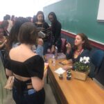 Autora sentada em mesa de autógrafos conversa com estudantes em evento acadêmico, com livros expostos ao seu lado.