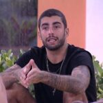 Homem de cabelo curto e barba, com tatuagens nos braços, falando com outras pessoas enquanto está sentado em ambiente interno com plantas ao fundo