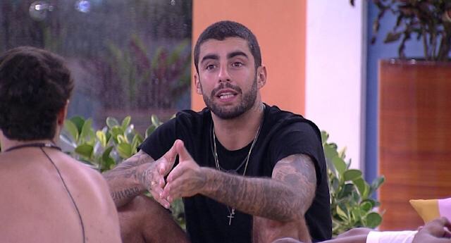 Homem de cabelo curto e barba, com tatuagens nos braços, falando com outras pessoas enquanto está sentado em ambiente interno com plantas ao fundo