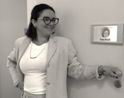 Psicóloga Gisa Dineli sorrindo em frente à porta de seu consultório, com placa de identificação ao lado. Psicoterapia para Expatriados